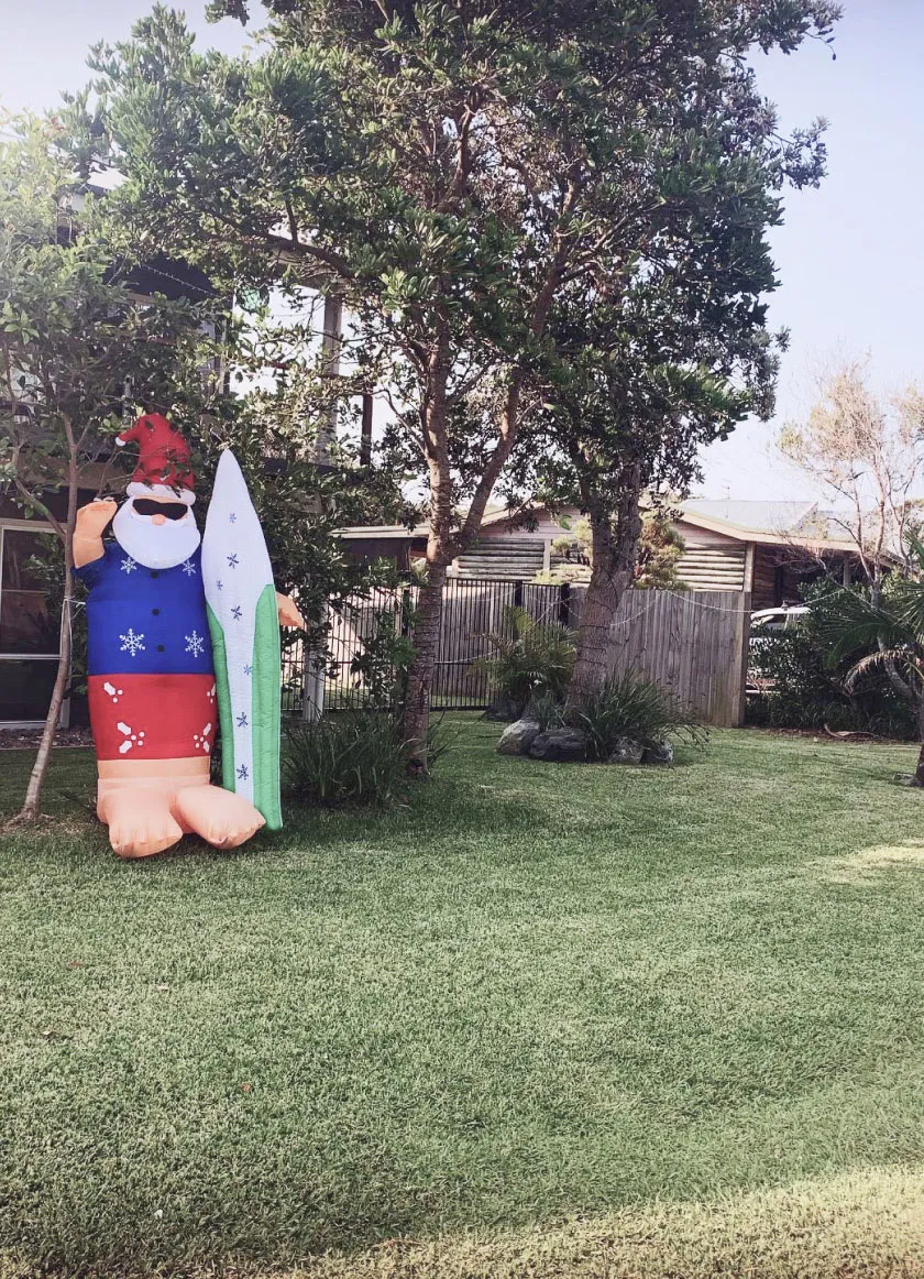 An inflatable Santa Claus with a surfboard in a suburban yard, celebrating a beach-themed Christmas