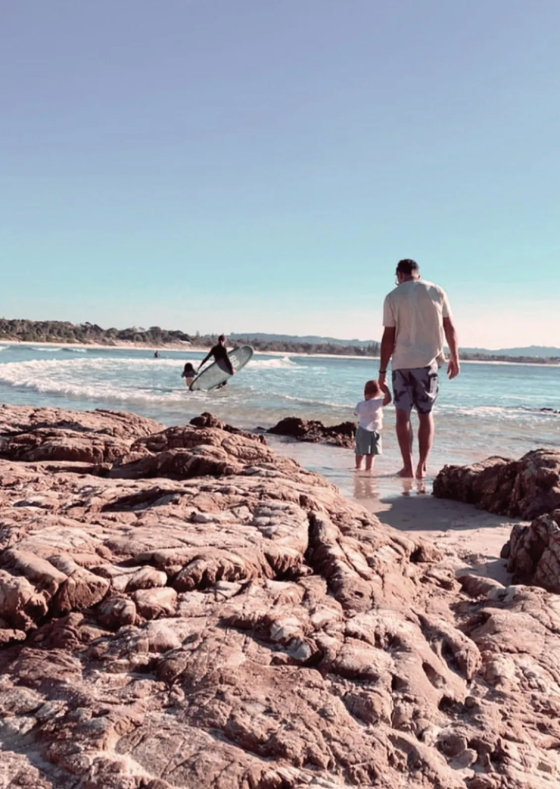 Antonio and his son walking hand in hand on a rocky beach shore, with another person surfing in the background